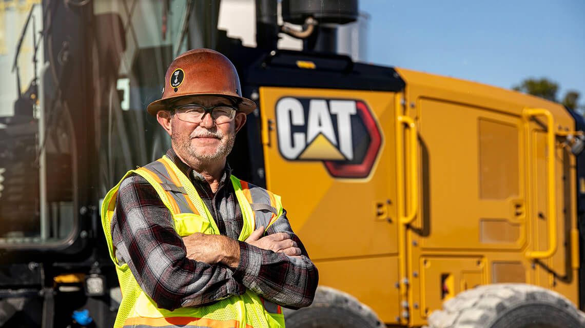 Construction worker wearing a hard hat crossing his arms in front of a Cat machine